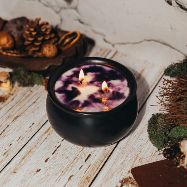 Candle in a black holder with purple wax, surrounded by dried flowers and pinecones on a wooden surface.