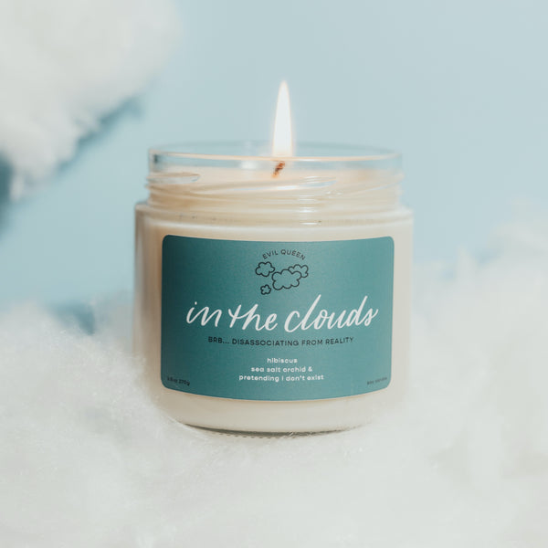 A white candle with a blue label that reads 'in the clouds' with a cloud illustration, placed on a surface with a blue out-of-focus background.